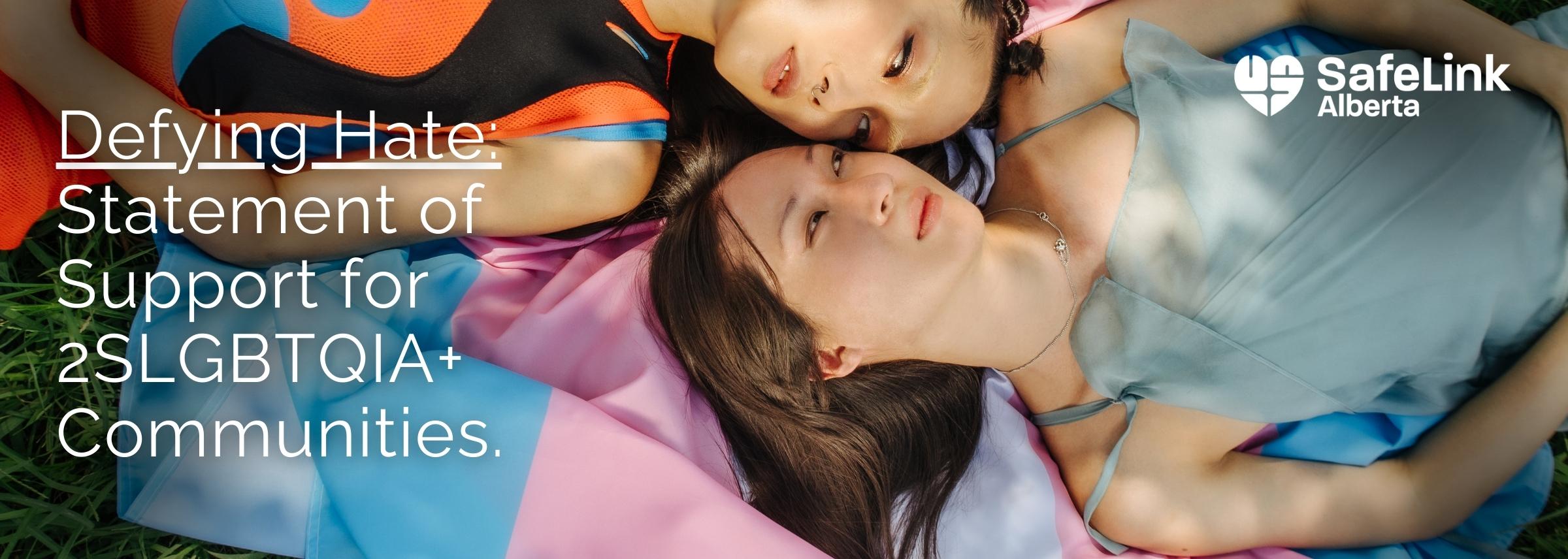 Text Reads "Defying Hate: Statement of Support for 2SLGBTQIA+ Communities" Image shows two trans people laying on a trans flag in the grass, dappled with sunlight.