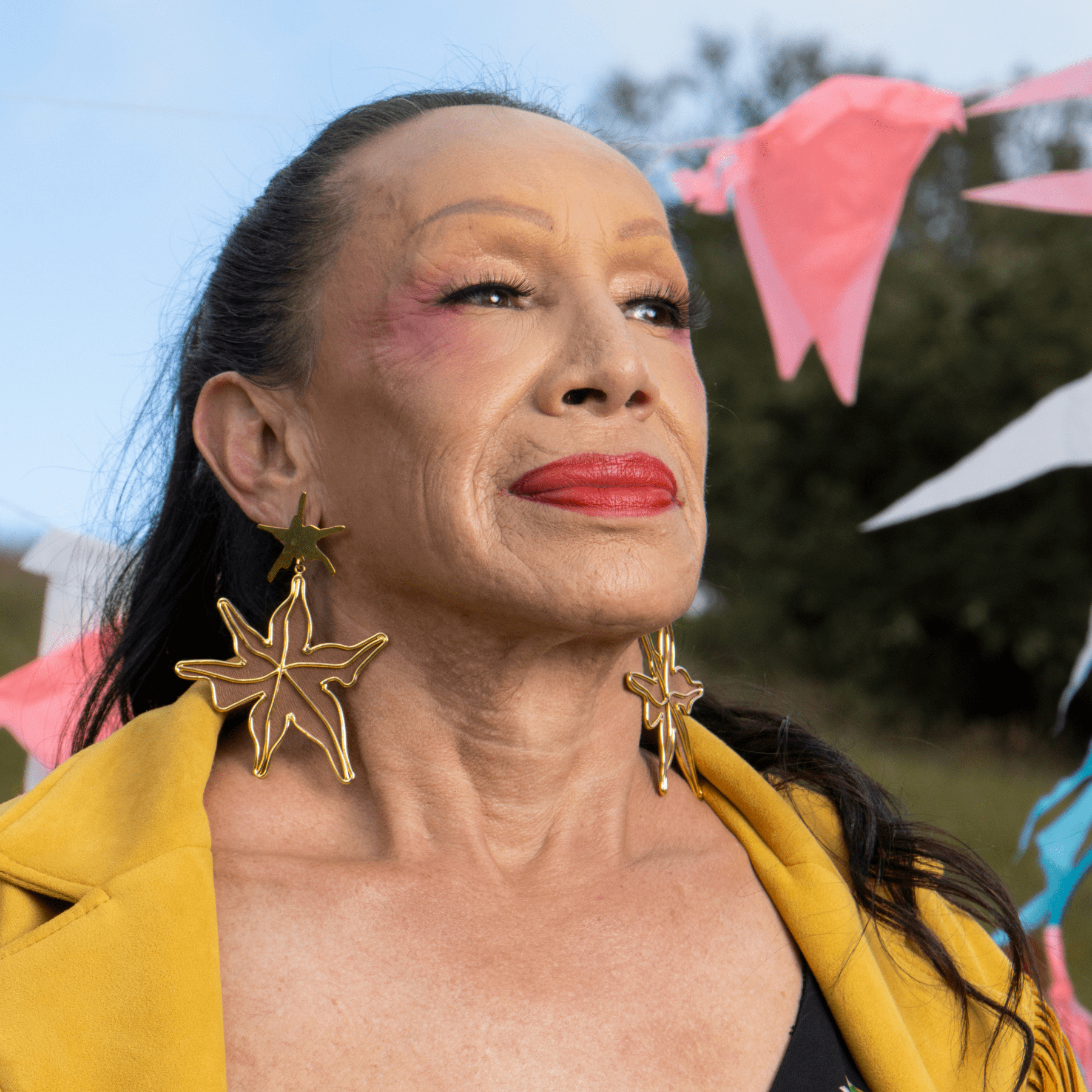 Image of an older woman wearing red lipstick, large gold starfish earrings, and yellow jacket.