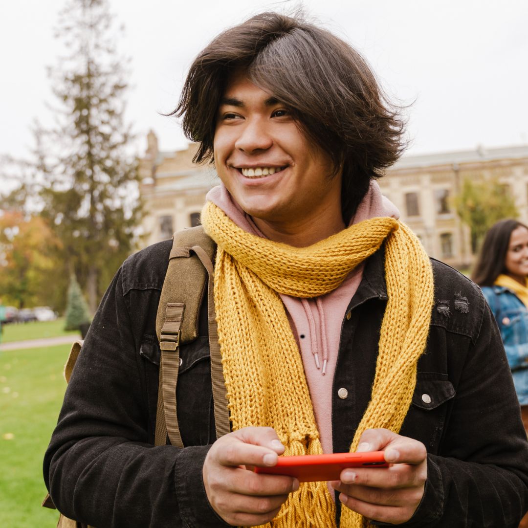 Young, handsome, indigenous man, on a school campus with a backpack. Smiling and holding a phone. 