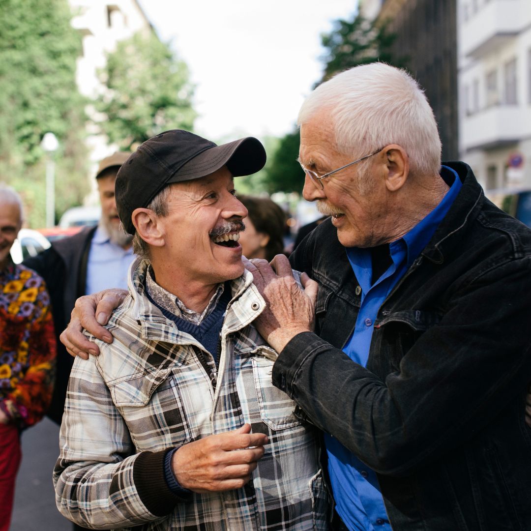Two older white gay men embrace each other,  smiling into each other eyes. Their friends are walking down a street with them. 