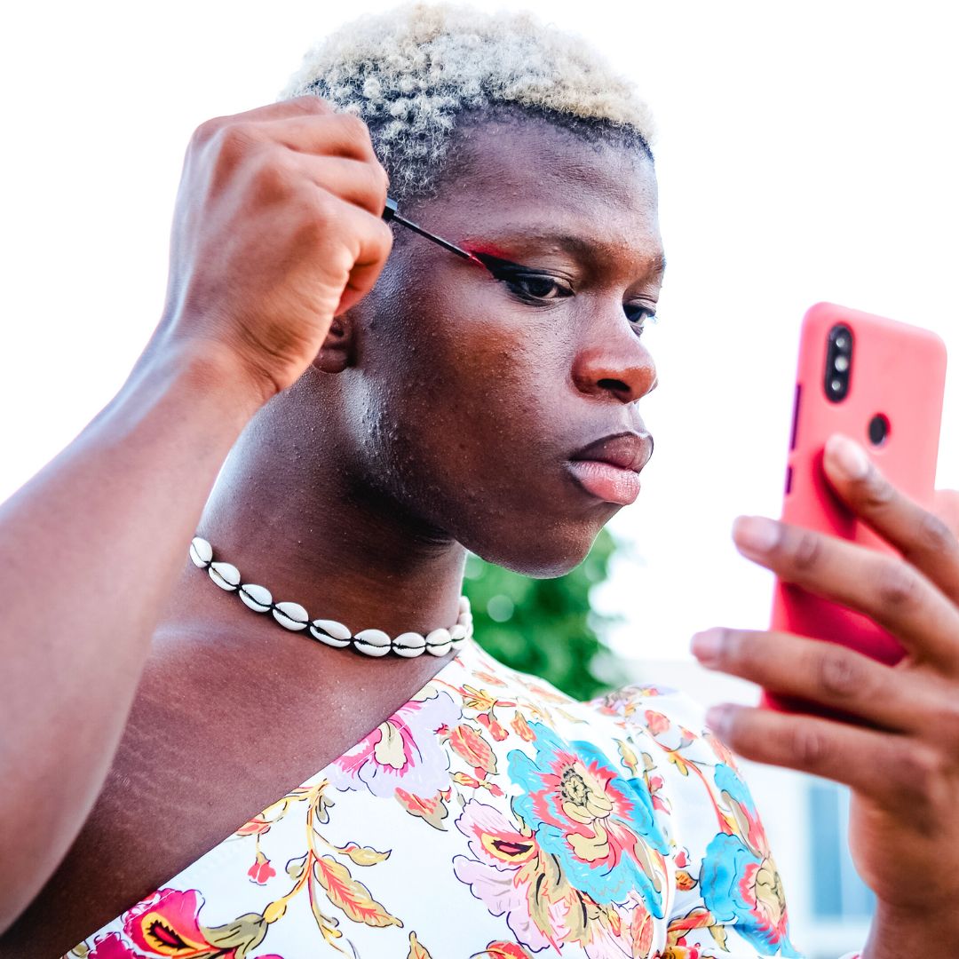 A young african american non-binary person, wearing a dress and applying makeup with their phone camera.