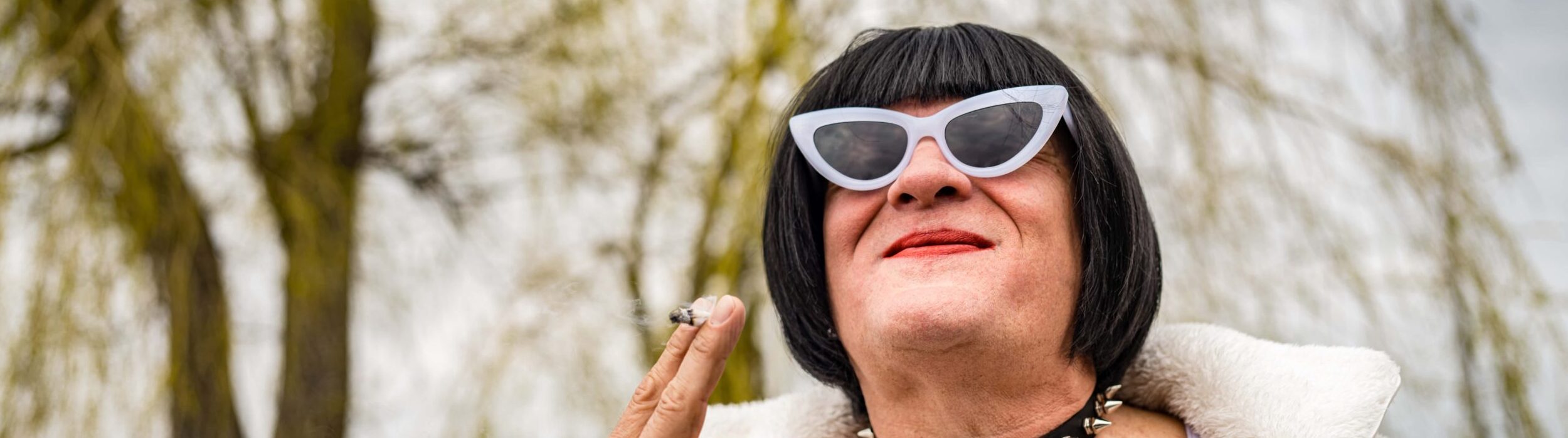 Transgender woman with cool sunglasses, a white coat, a black bob haircut. She is holding a cigarette. 