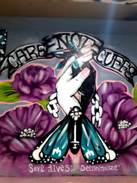 A mural reading "Care Not Cuffs" in Medicine Hat Alberta. It depicts two hands holding each other, with purple flowers surrounding them. The bottom text reads "Save Lives Decriminalize". This mural was created to mark International Overdose Awareness Day.