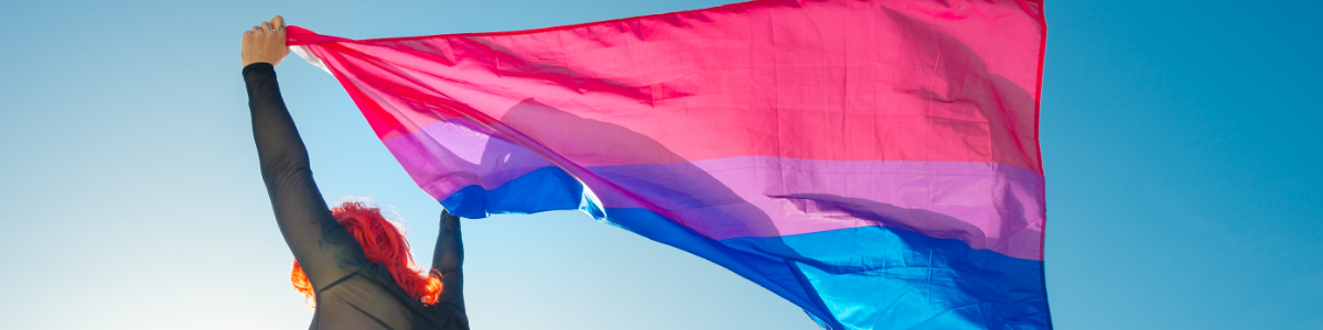 Woman with red hair holding a bi flag against a clear blue sky.