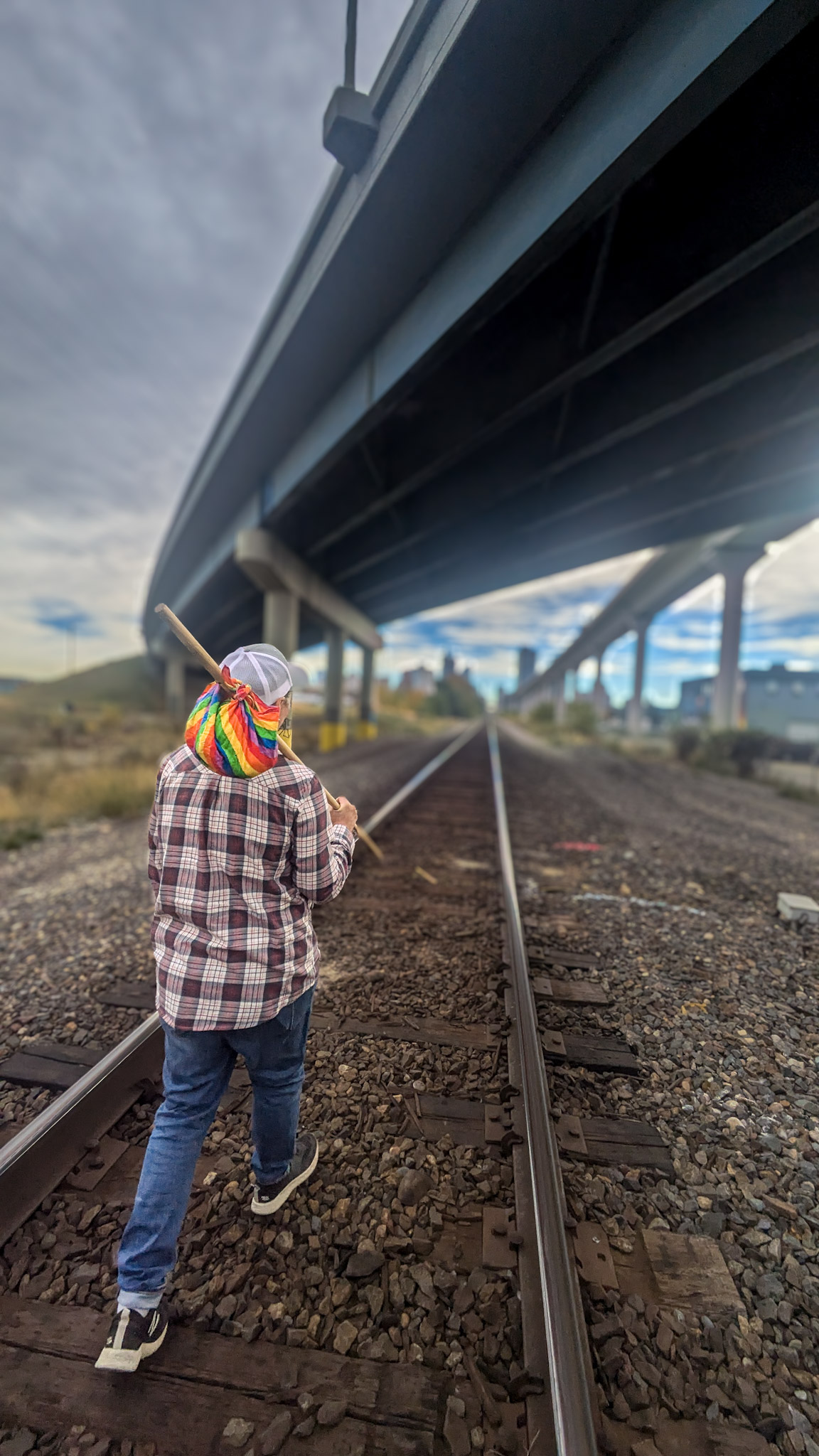 An image of a person carrying a rainbow coloured bindle down a railroad track. Meant to symbolize the risk of homelessness that comes with coming out as 2SLGBTQIA+. 